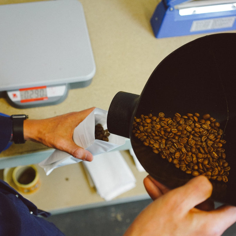 Person transferring coffee beans from a scoop into a paper bag on a scale.